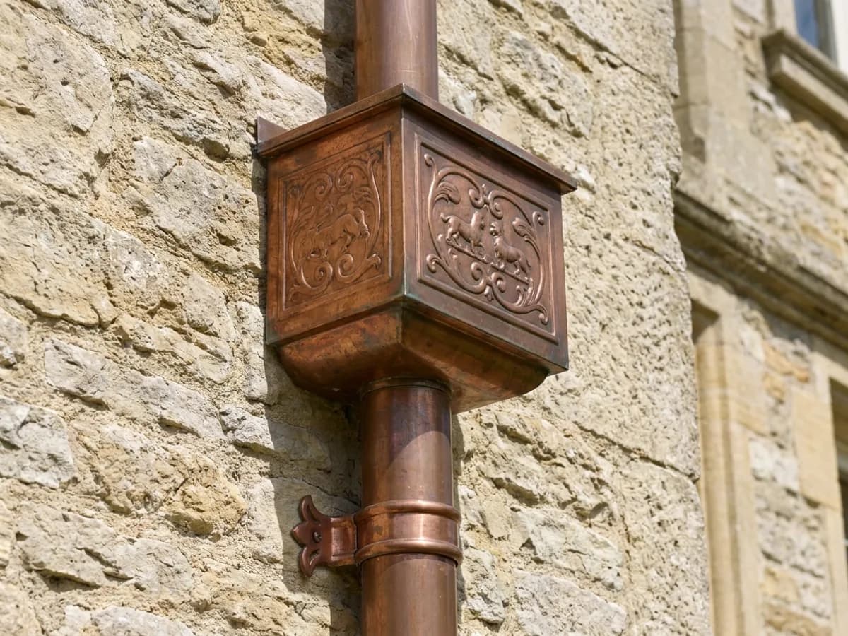 Ornate copper leader head and round downspout on a limestone wall